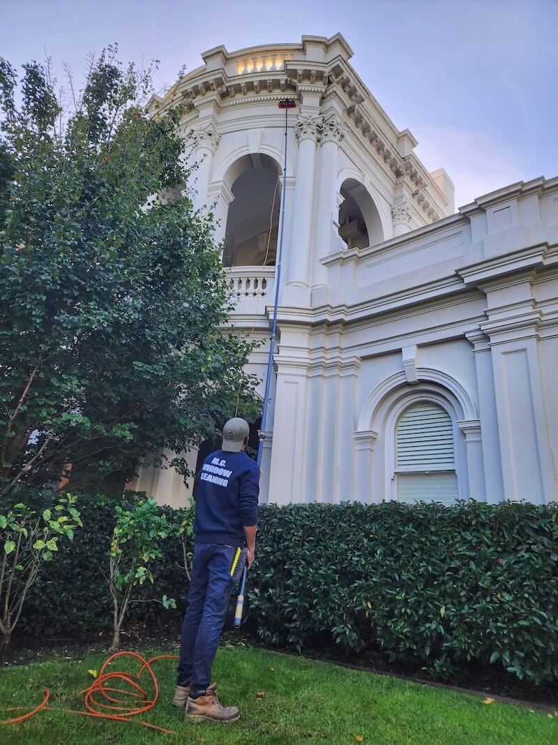 Person wearing MC Window Cleaning uniform using an extendable pole to clean the upper windows of a white ornate building.