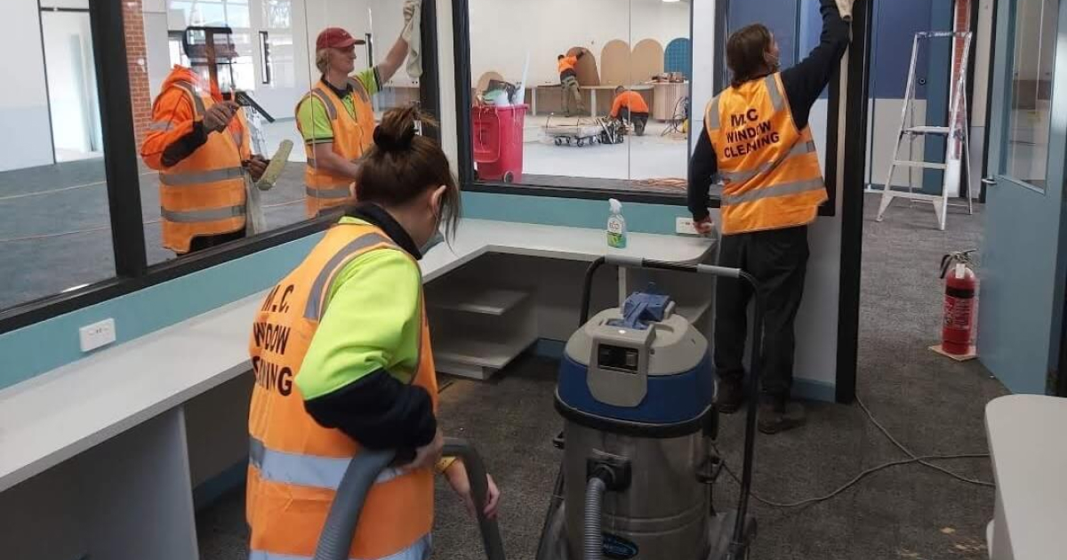Four workers wearing orange safety vests cleaning large glass windows and vacuuming inside an office space under renovation.