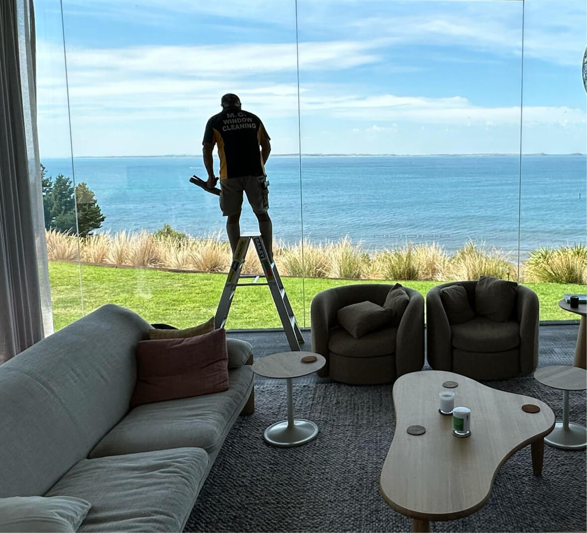 Man on ladder cleaning large glass window overlooking a grassy yard and ocean view.