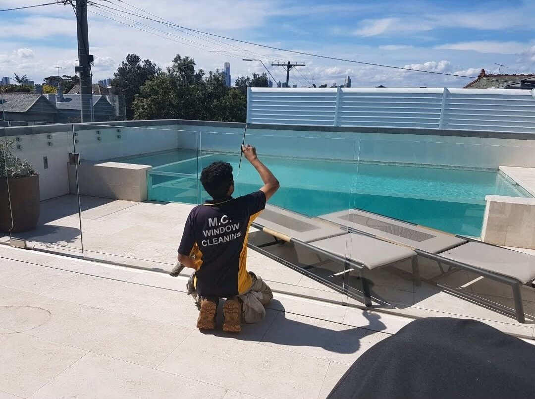 A worker in an M.C. Window Cleanings shirt cleans glass panels surrounding a pool on a rooftop terrace under a blue sky.